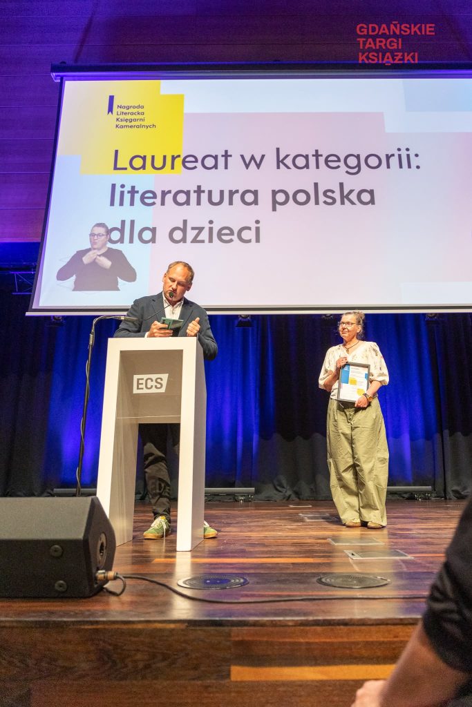 Przemówienie przy mównicy podczas gali w&nbsp;Gdańsku, w&nbsp;tle laureatka Jola Richter-Magnuszewska słucha laudacji z&nbsp;dyplomem w&nbsp;dłoniach.
